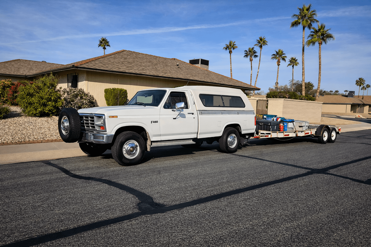 White vintage Ford F-250 with camper shell towing a loaded trailer past palm trees.